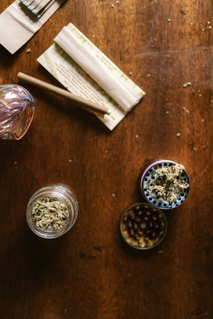 Glass jar, grinder, and joint papers on a wooden table, representing cannabis culture.